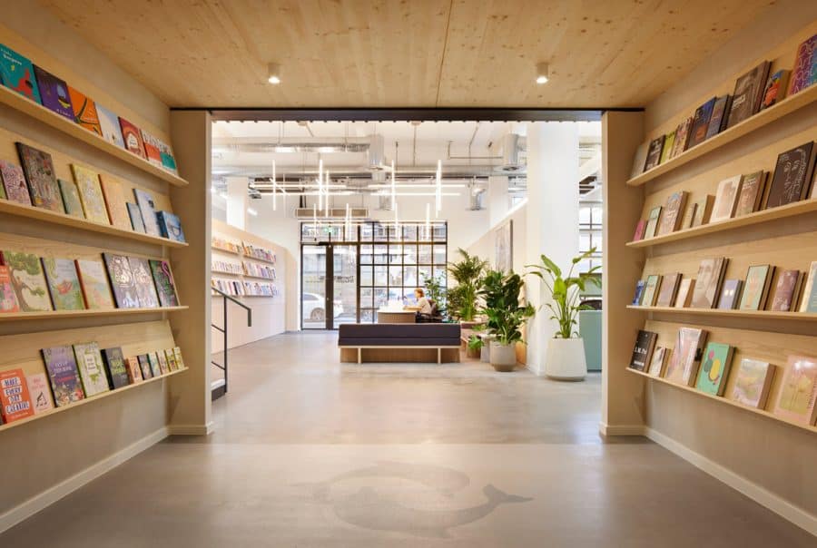 Interior of a modern publishers office with wooden shelves filled with books on both sides. A receptionist sits at a desk in a seating area surrounded by large potted plants. The space is well-lit with hanging contemporary lights and natural light from large windows.