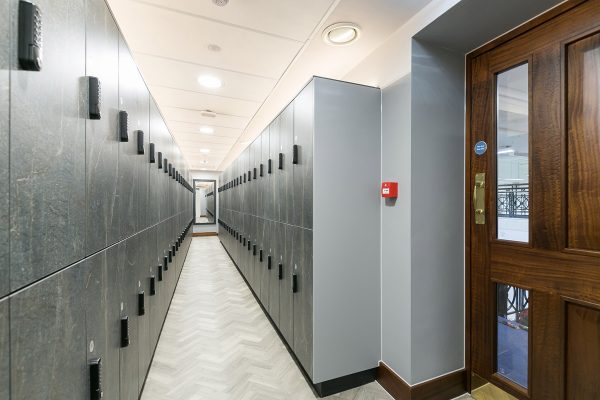 Refurbished locker lined locker room with modern grey lockers, enhanced with herringbone flooring, updated lighting, and safety features like a fire alarm—blending functionality with a clean, contemporary aesthetic.