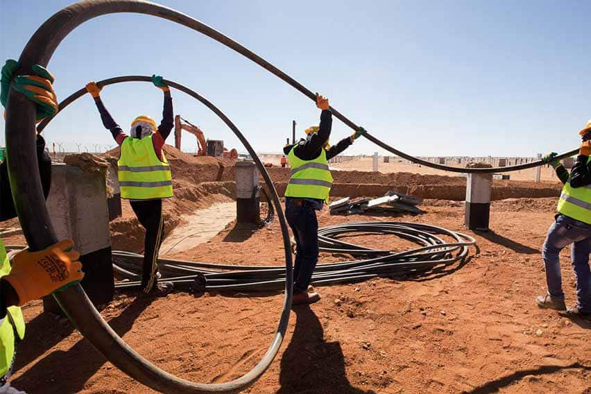 Three construction workers in yellow safety vests and helmets handling a large black cable at a construction site with reddish dirt, concrete pillars, and construction equipment in the background.