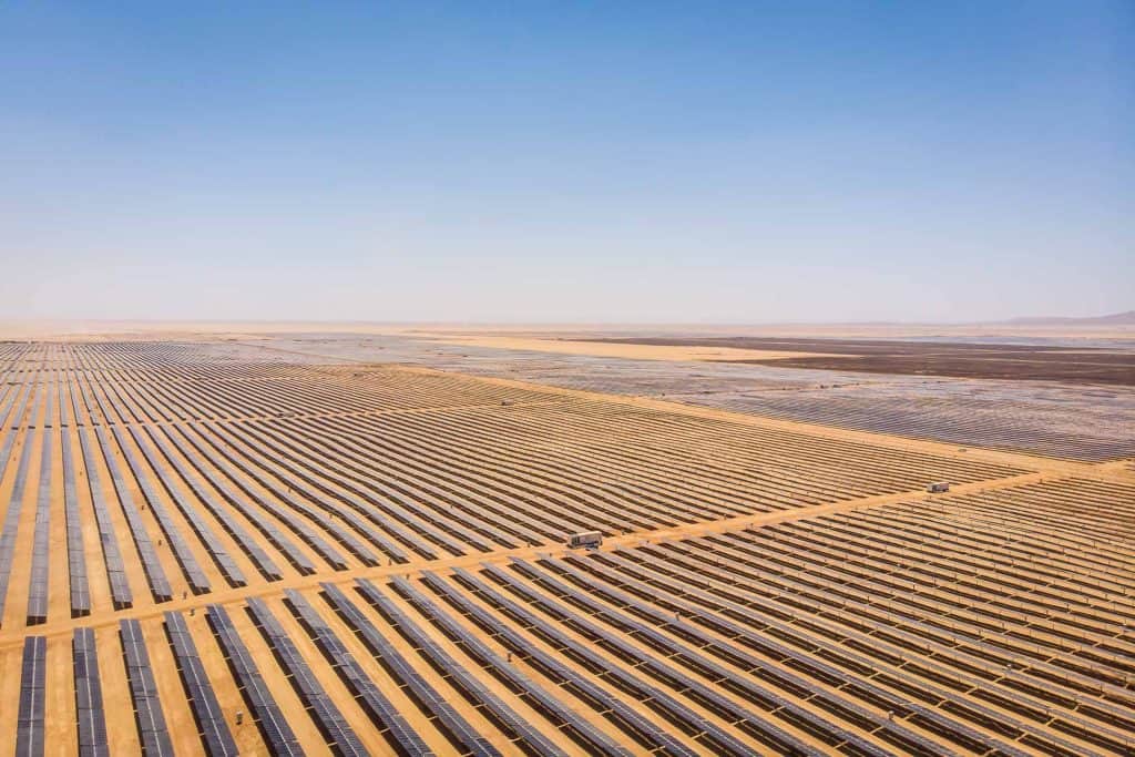 A vast solar farm with rows of solar panels arranged in a grid pattern across a flat, arid landscape under clear skies.