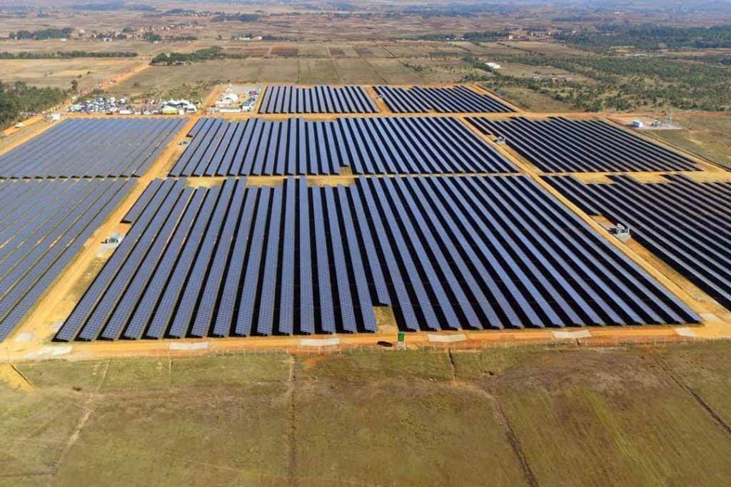 A large solar farm with rows of solar panels arranged in parallel lines across a wide rural area; buildings and structures in the background suggest maintenance or operational facilities, with open fields and sparse vegetation surrounding the site.