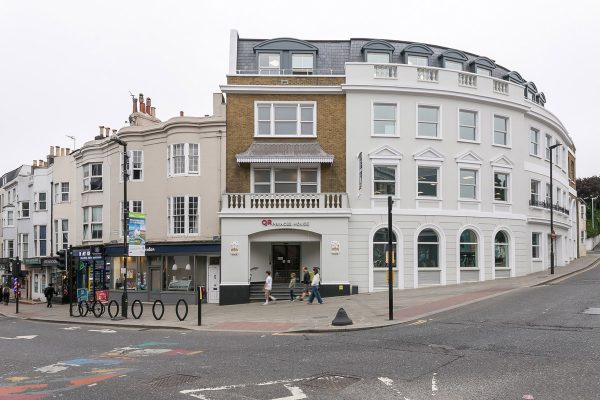Street corner with a white building featuring a curved facade and multiple windows in Brighton. People are walking on the sidewalk, with bollards and a lamppost nearby. The street has colorful markings, possibly indicating a pedestrian crossing or bike lane.