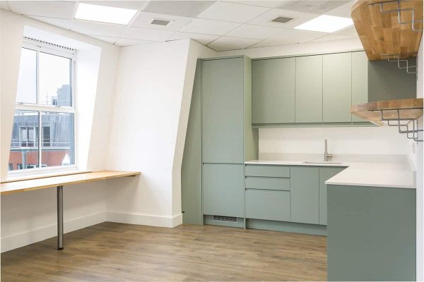 Modern office kitchenette with sage green cabinets, white countertops, a built-in sink, and wooden flooring, featuring a large window with a wooden desk beneath and floating wooden shelves on the wall.