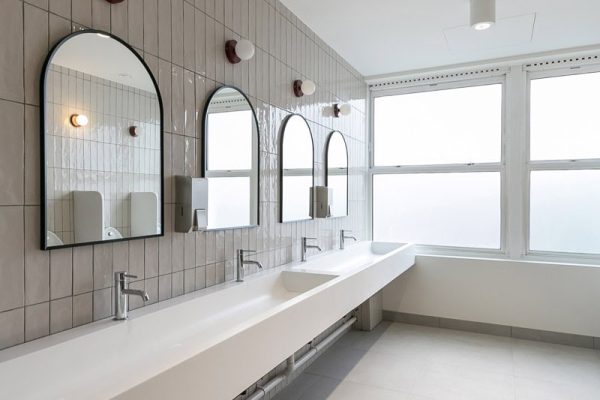 Bright restroom with a long white trough sink, multiple chrome faucets, arched mirrors, wall-mounted soap dispensers, and large frosted windows allowing natural light to enter