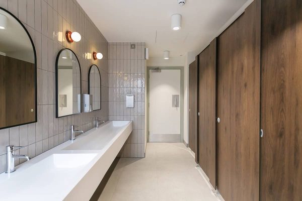 Bright restroom with a long white trough sink, multiple chrome faucets, arched mirrors