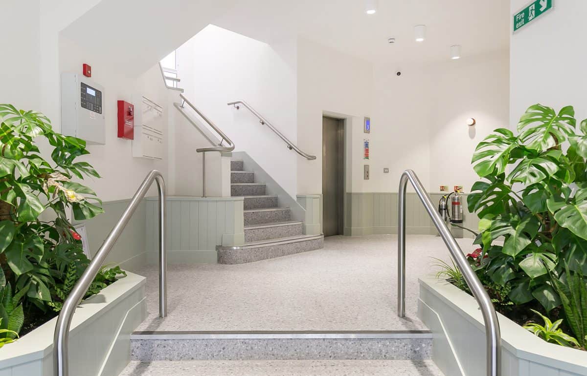 Indoor space with a staircase leading to an upper floor, flanked by handrails and two large green plants. To the right of the stairs is an elevator with control buttons and indicators. The left wall features a fire alarm system and a red fire extinguisher. The area has white walls, light-colored flooring, and is brightly lit.