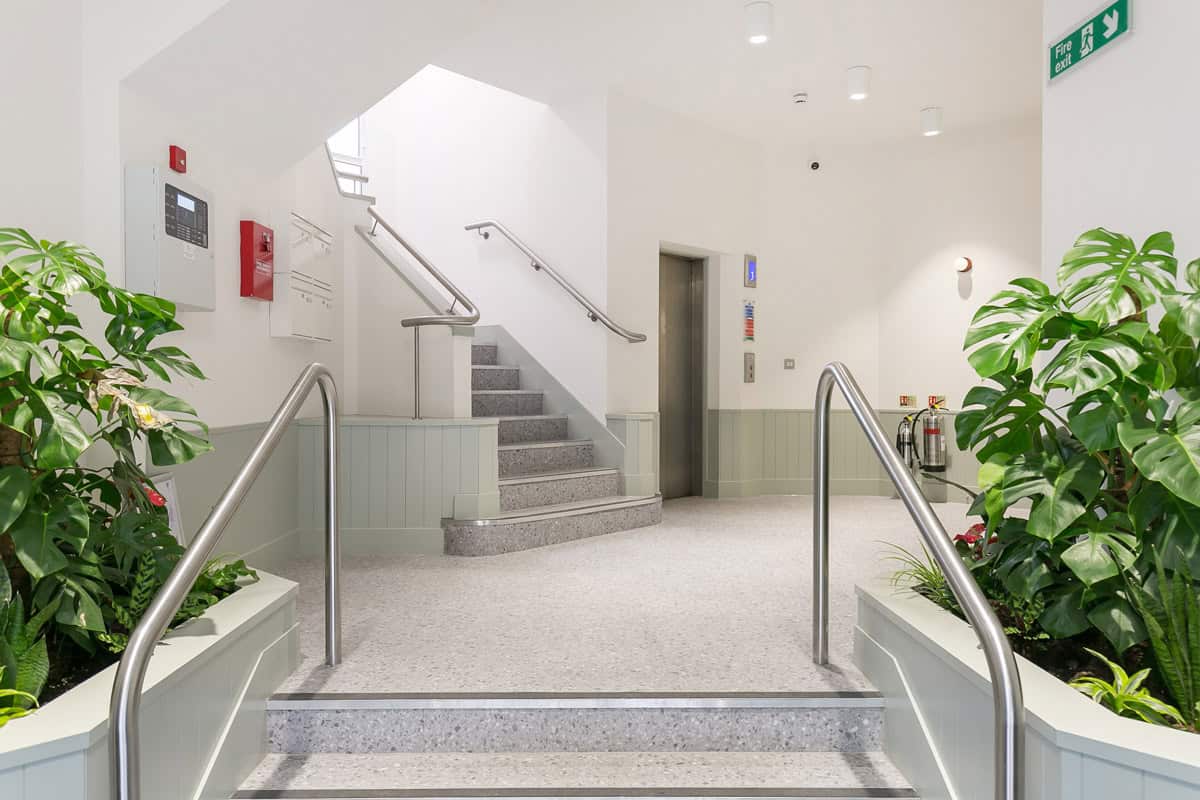 Indoor space with a staircase leading to an upper floor, flanked by handrails and two large green plants. To the right of the stairs is an elevator with control buttons and indicators. The left wall features a fire alarm system and a red fire extinguisher. The area has white walls, light-colored flooring, and is brightly lit.