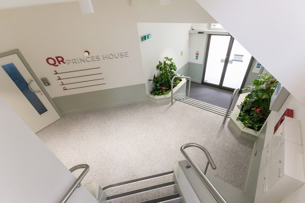 Bright and clean building entrance with stairs leading to glass double doors, indoor plants in built-in planters, and wall signage reading 'QR Princes House' with floor directory indicators
