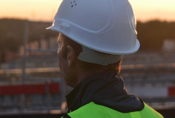 A construction worker wearing a white hard hat and a bright yellow safety vest, seen from behind, looking out over a construction site during sunset.