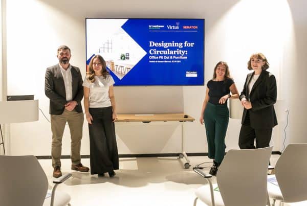 Four individuals standing in front of a large screen in a bright, minimalist room. The screen shows a slide titled “Designing for Circularity: Office Fit Out & Furniture” with logos for Broadbase, Virtus, and Senator. Empty chairs are visible in the foreground.