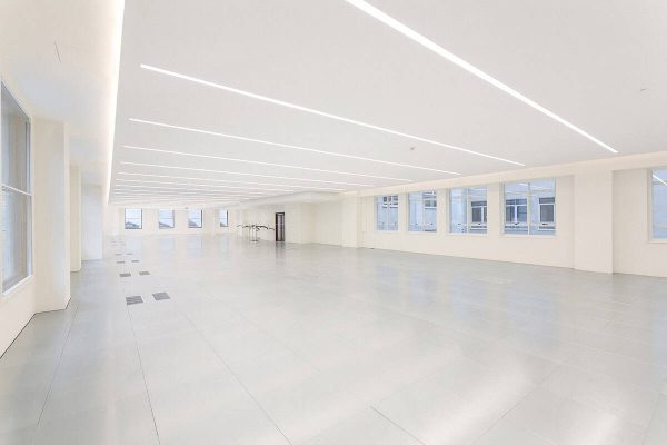 1-2 Bright, open-plan office space with white walls, large windows, and sleek underfloor air conditioning vents, featuring integrated linear LED lighting across the ceiling.