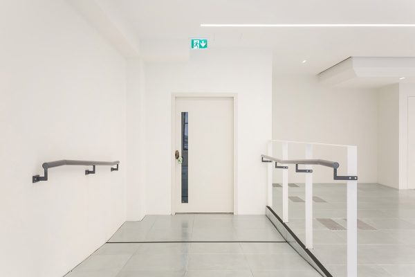 1-25 Minimalist office entrance area featuring a white door with a vertical glass panel, sleek handrails on both sides, and a light grey tiled floor, illuminated by integrated linear ceiling lights.