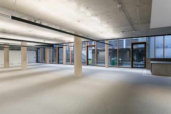 Spacious open office floor with concrete ceiling, structural columns, and full-height windows. Neutral carpet flooring and integrated floor boxes for power. Refurbishment could include partitioning options and acoustic treatment.