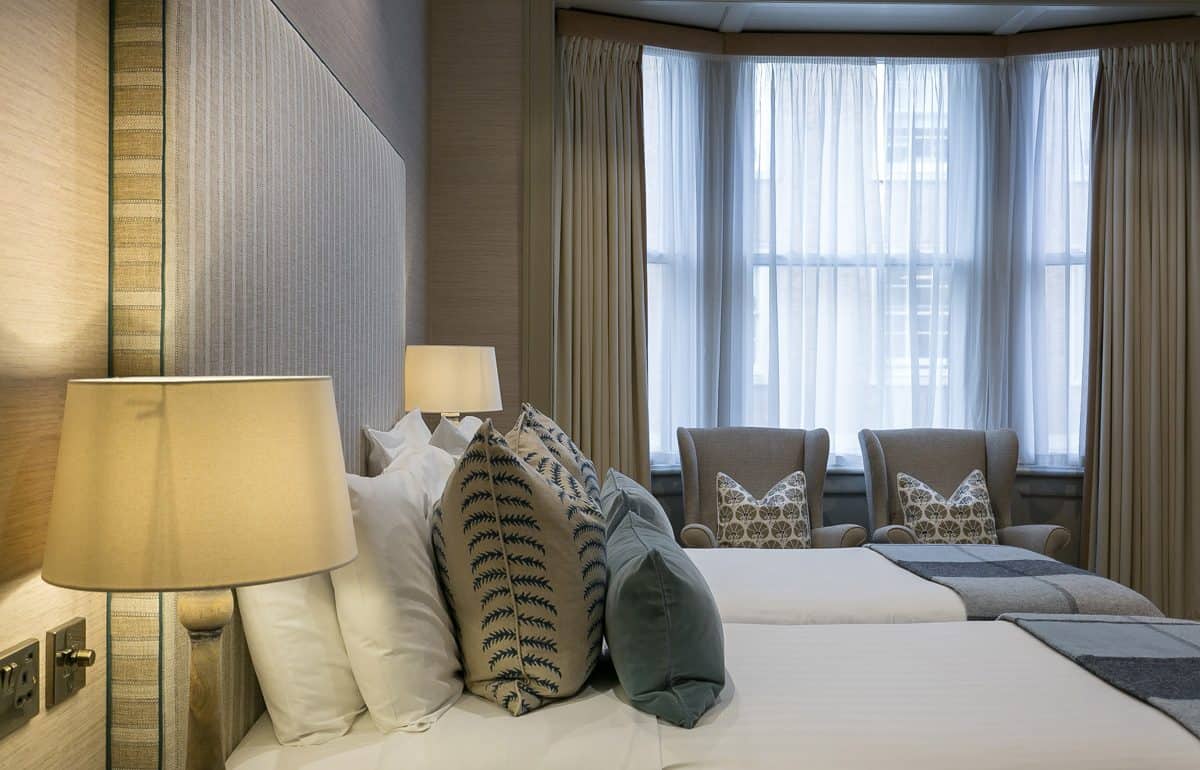 Twin beds positioned near a large bay window with sheer curtains and two upholstered armchairs. Warm beige tones dominate the space. Refurbishment could enhance natural light and add blackout options for improved comfort.