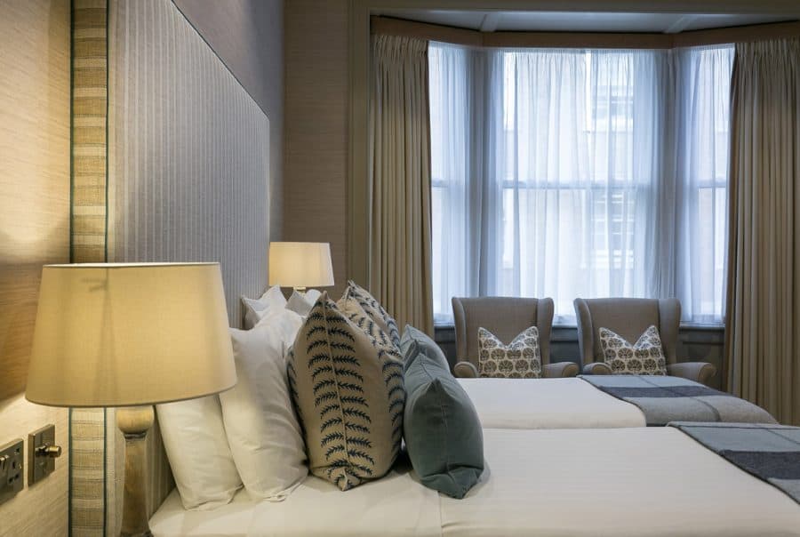 Twin beds positioned near a large bay window with sheer curtains and two upholstered armchairs. Warm beige tones dominate the space. Refurbishment could enhance natural light and add blackout options for improved comfort.