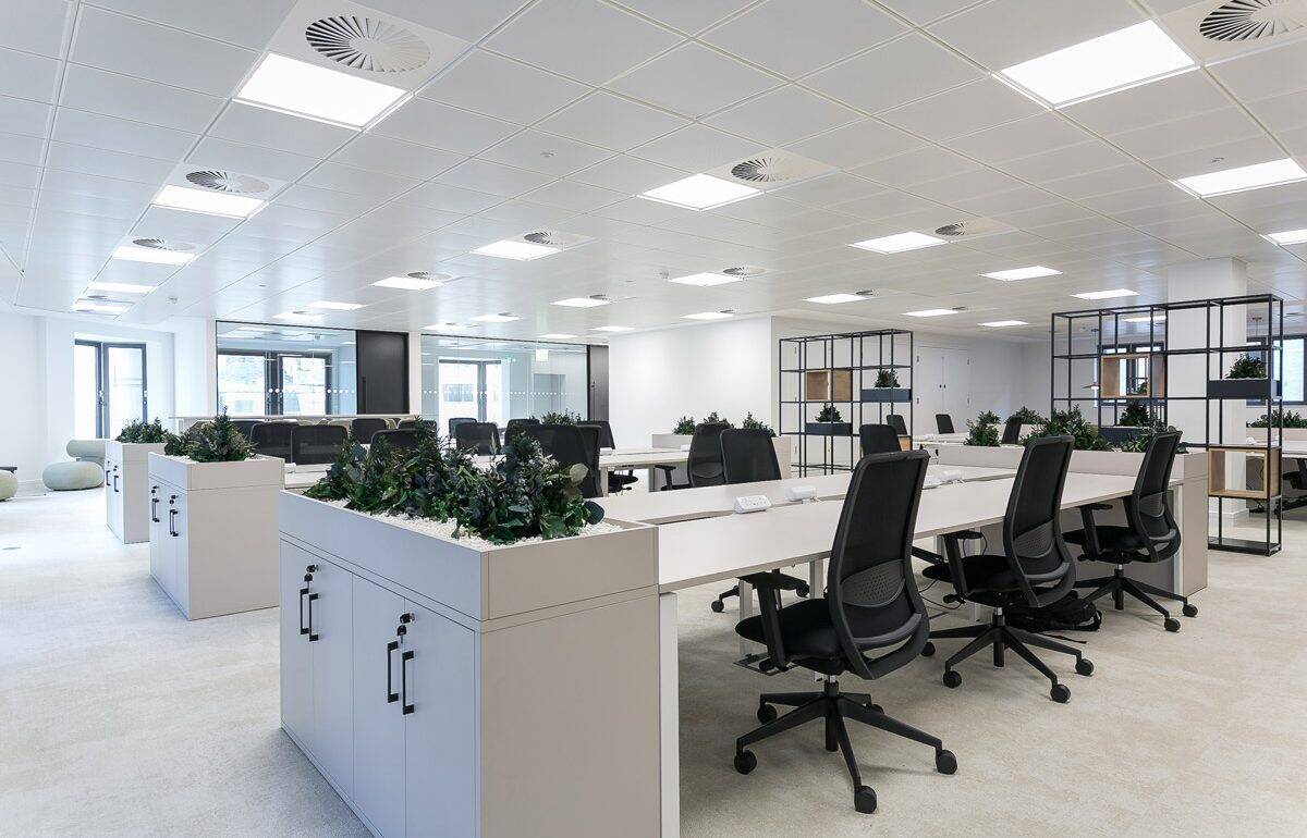 Spacious open-plan office with white desks, black chairs, and planter-topped storage units. Ceiling grid with LED panels and air diffusers ensures optimal lighting and ventilation.