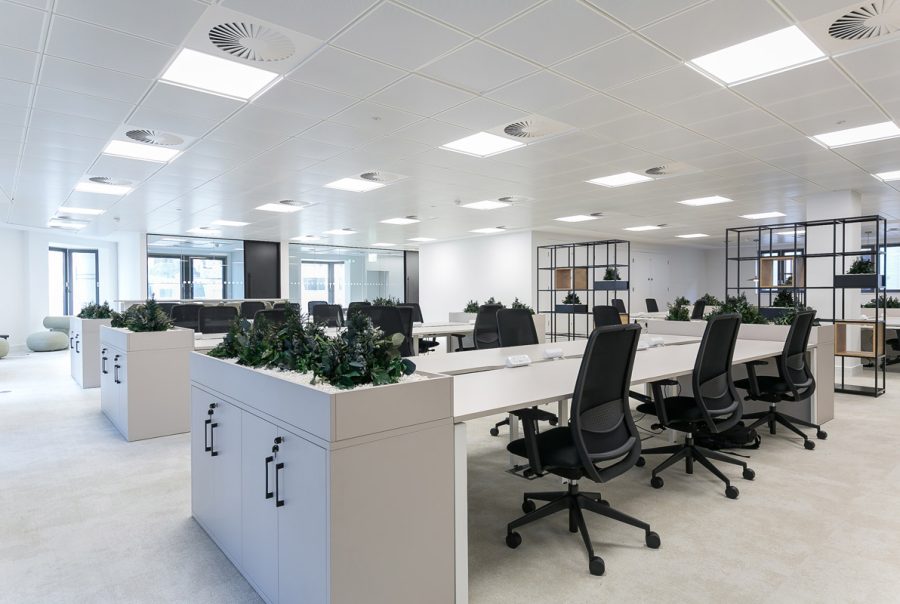 Spacious open-plan office with white desks, black chairs, and planter-topped storage units. Ceiling grid with LED panels and air diffusers ensures optimal lighting and ventilation.