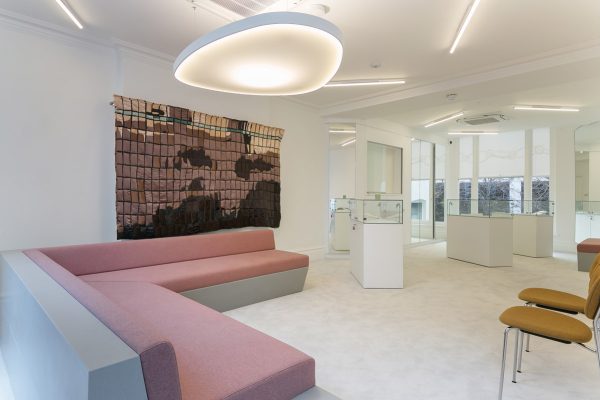 Wide-angle view of the refurbished showroom featuring bespoke seating, glass display units, and integrated lighting solutions. The design combines soft furnishings with hard finishes for a balanced, modern aesthetic. Pendant lighting and mirrors enhance brightness and spatial perception.