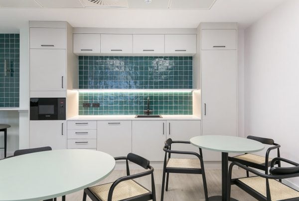 Compact kitchen and dining area featuring teal tiled splashback, white cabinetry, and integrated lighting. Round tables and black-framed chairs create a modern, functional breakout space.