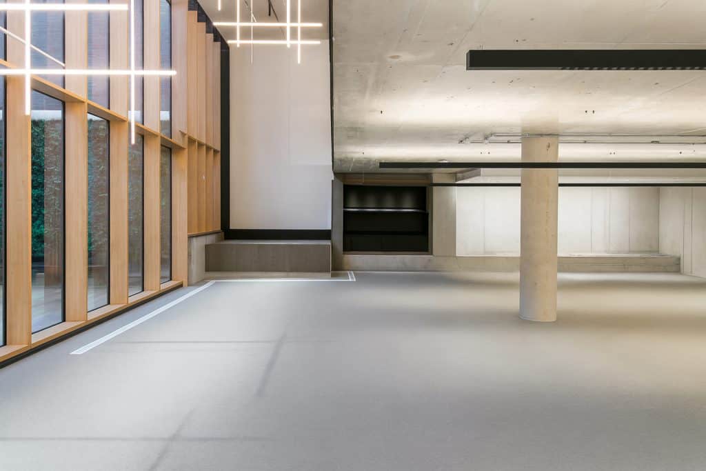 Minimalist office corner with large windows, concrete ceiling, and integrated shelving. Linear pendant lighting and neutral flooring dominate the space. Refurbishment could add collaborative zones and soft furnishings for comfort.