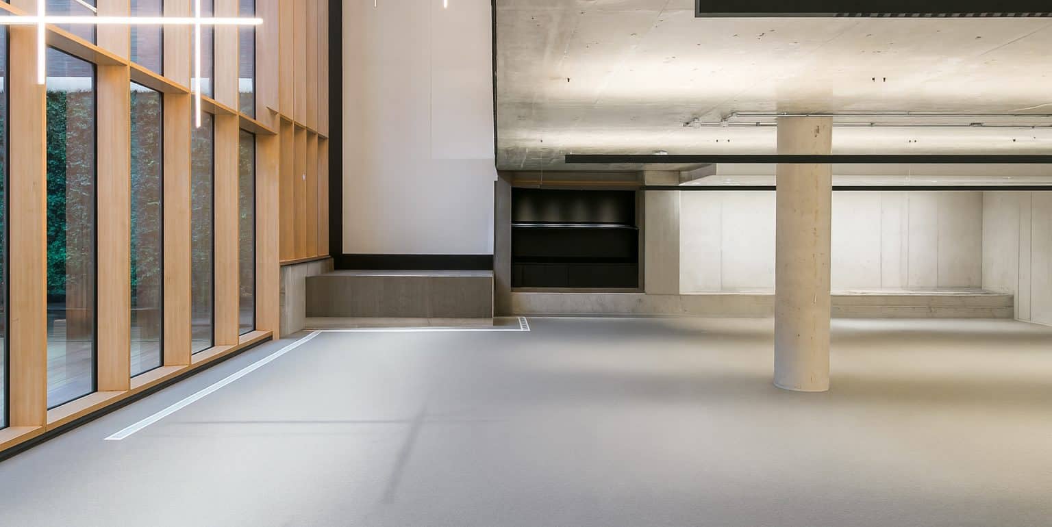 Minimalist office corner with large windows, concrete ceiling, and integrated shelving. Linear pendant lighting and neutral flooring dominate the space. Refurbishment could add collaborative zones and soft furnishings for comfort.