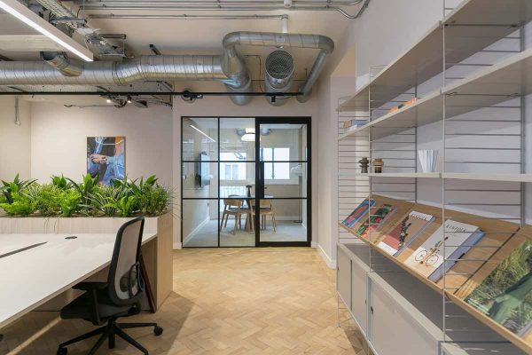 A workspace with long desks, greenery-filled planter dividers, and a shelving unit displaying magazines. A glass meeting room is visible in the background, with exposed ceiling ducts overhead.