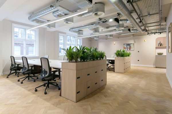 A bright office space with long shared desks, planter boxes dividing work areas, herringbone wood floors, and large windows letting in natural light. Exposed ceiling ducts create an industrial aesthetic.