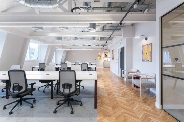 An office workspace with white desks and ergonomic chairs placed on carpet, with herringbone wood flooring around the perimeter. Exposed ceiling ducts and wall art add to the modern design.