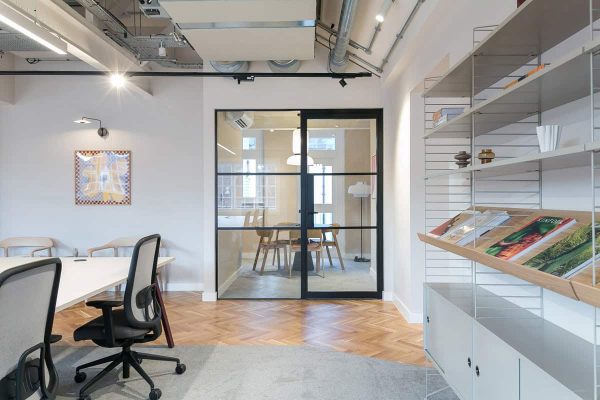 A view of an office space featuring a large glass meeting-room door, shelving with magazines, and part of a long communal desk with task chairs. Light wood floors and exposed ceilings add an industrial style.