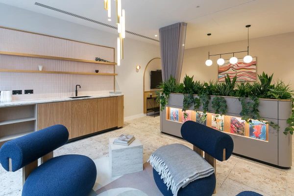 A lounge-style space with modern blue chairs, a small marble table, a kitchenette with open shelving, and a planter divider with integrated lighting. Soft colors and plants create a warm atmosphere.