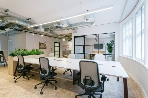 A curved desk area with several office chairs, herringbone floors, and a large glass meeting-room door. Exposed ducts run along the ceiling, and a tall plant sits near the window.