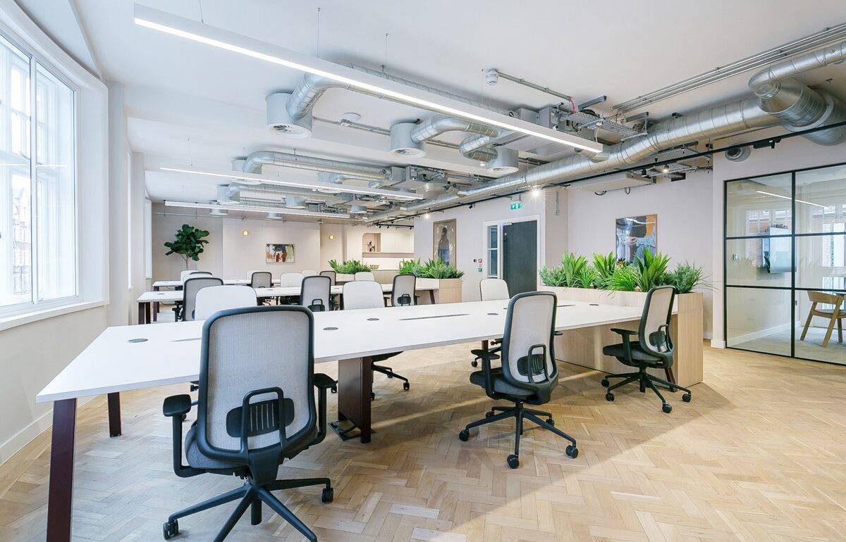 A bright open-plan office with long white desks, ergonomic chairs, planter dividers, herringbone floors, and exposed ceiling ducts. A glass meeting room and several artworks line the walls.