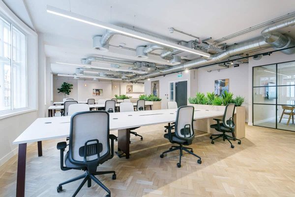 A bright open-plan office with long white desks, ergonomic chairs, planter dividers, herringbone floors, and exposed ceiling ducts. A glass meeting room and several artworks line the walls.
