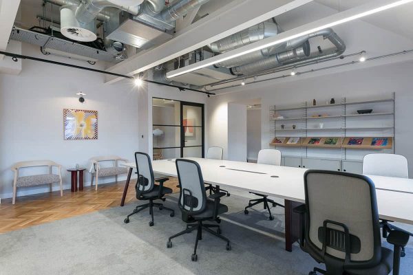 A modern open-plan office with a long white desk, several ergonomic chairs, exposed ceiling ducts, and shelves holding books and decorative objects. Light wood flooring transitions into carpet around the desks.