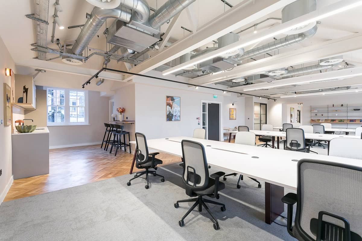 A large office with long desks and office chairs, greenery planters, herringbone floors, and exposed overhead ductwork. A bar-height table and stools sit near a window in the back corner.