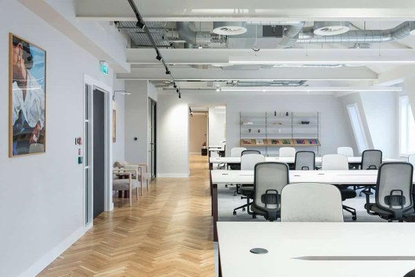 An open office area with rows of white desks and office chairs, light wood herringbone floors, exposed ducts on the ceiling, and wall-mounted shelving holding books and decorative pieces.