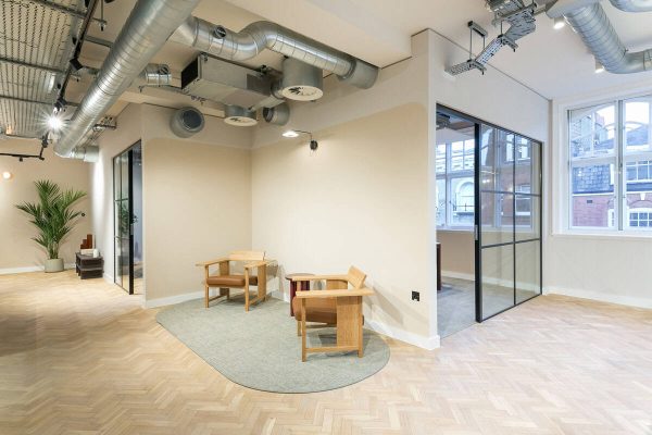 A small seating nook with two wooden lounge chairs and a circular rug, positioned under exposed ceiling ducts. A glass door leads to another office area, and artwork hangs on the wall.