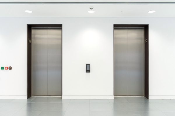 Refurbished lift lobby with two stainless steel elevators set into newly finished white walls, updated lighting, and modern grey floor tiles.
