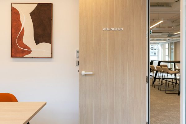 Interior office space with a light wood door labeled ‘ARLINGTON.’ To the left of the door is a modern abstract painting in earthy tones. A corner of a table and an orange chair are partially visible. Through the open door, a contemporary workspace with high stools and large windows can be seen.