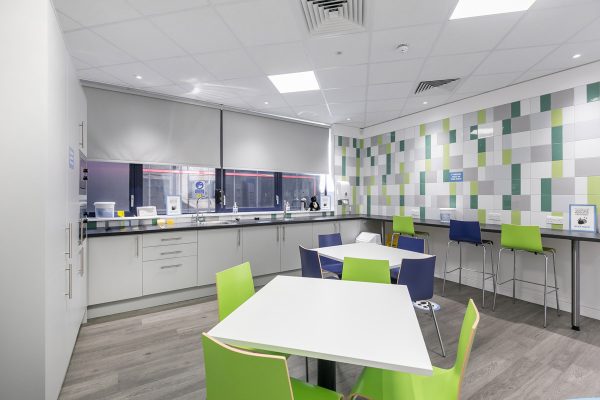 Staff kitchen and breakout space with tiled feature wall, fitted cabinetry, perimeter worktops and informal seating, delivered as part of CAT B office fit out.