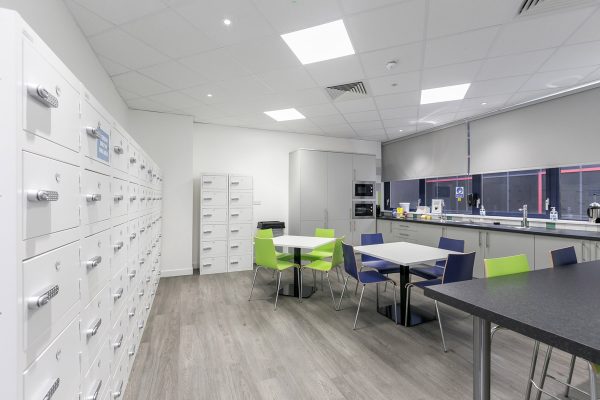 Breakout kitchen area within CAT B office fit out, featuring secure locker storage, integrated appliances, vinyl flooring and suspended ceiling with LED lighting and ventilation.